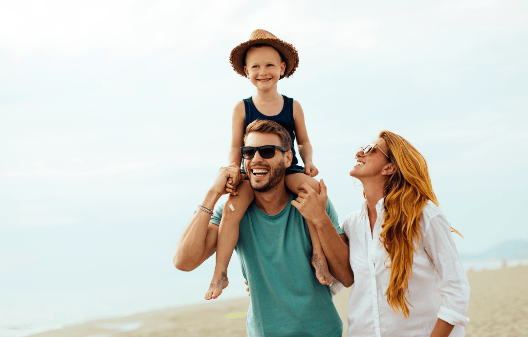 Joyful family at the beach with a child on shoulders, enjoying a sunny and affordable summer vacation.