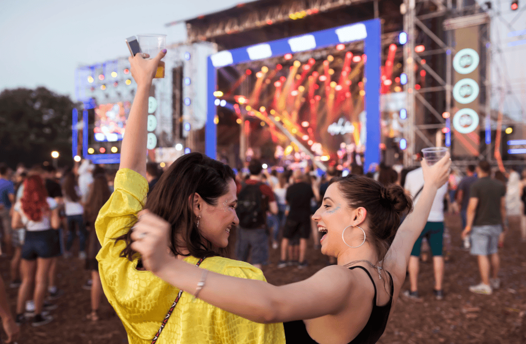 Two women dancing and celebrating at a summer music festival in Europe with stage lights and crowd