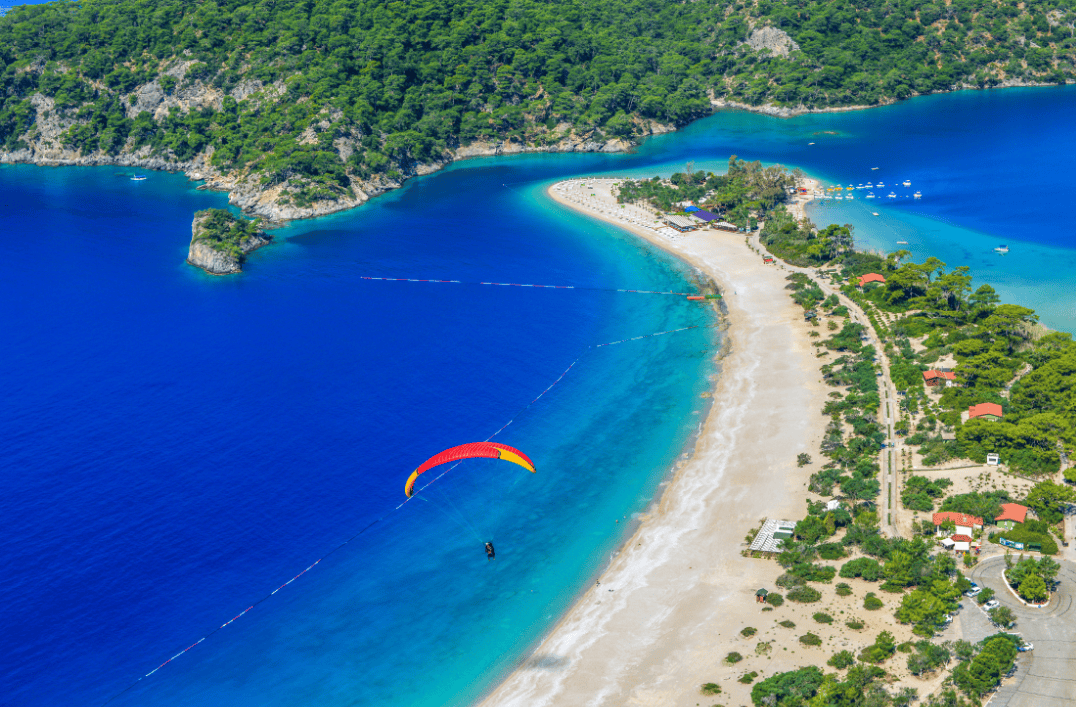 Aerial view of Oludeniz Beach in Fethiye, Turkey, with a paraglider soaring above turquoise waters and pine-covered coastline.