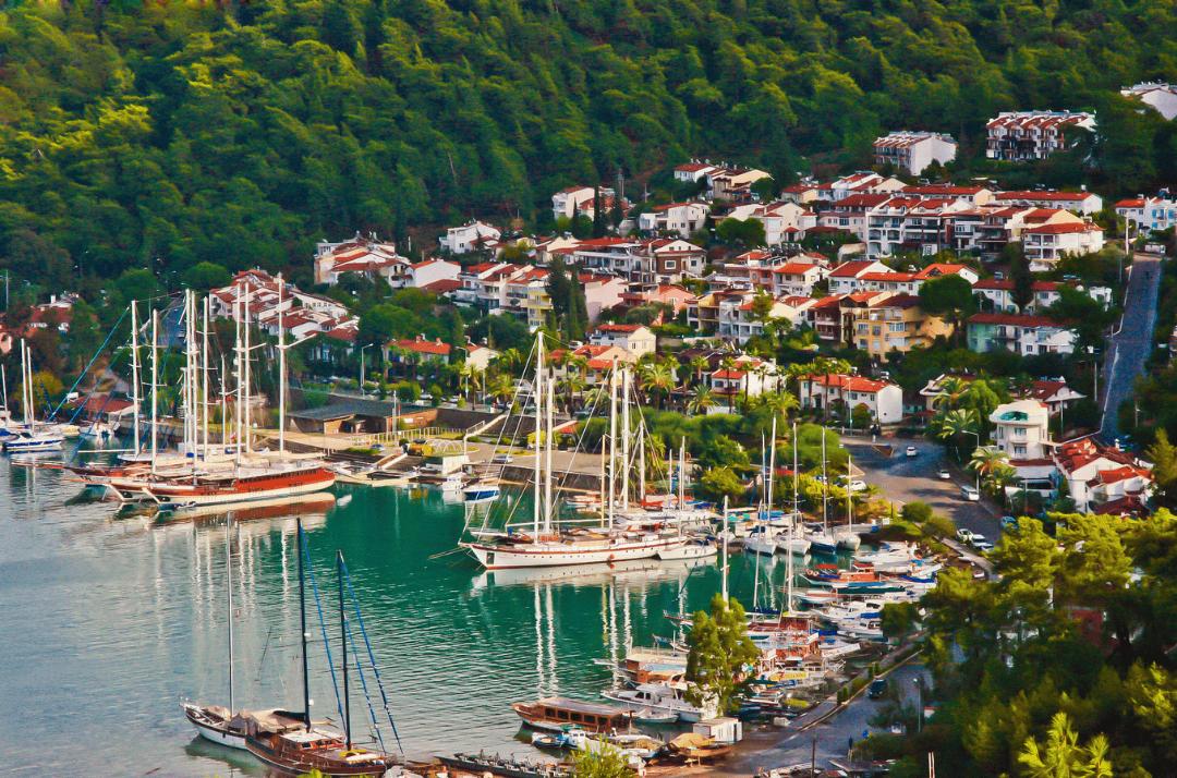 Scenic marina in Fethiye, Turkey, featuring moored sailboats, waterfront houses, and lush green hills in the background.