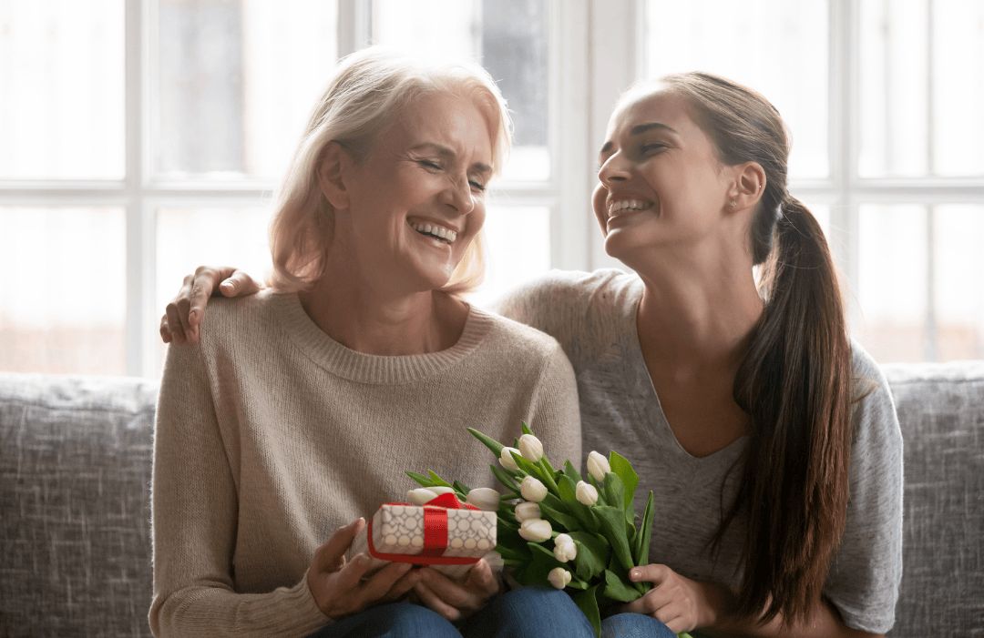 A happy mother and daughter sitting on a couch, smiling and holding a small gift box and a bouquet of white tulips.