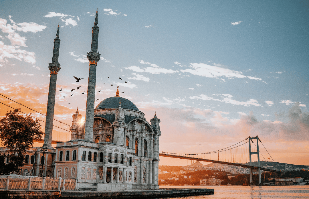 Ortaköy Mosque in Istanbul, Turkey, with birds flying at sunset and the Bosphorus Bridge stretching across the horizon.