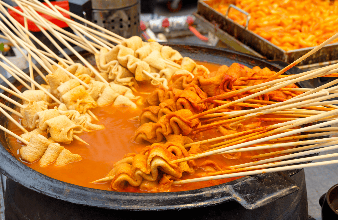 A large pot filled with skewered Korean fish cakes (odeng) simmering in hot, flavorful broth, with tteokbokki visible in the background.