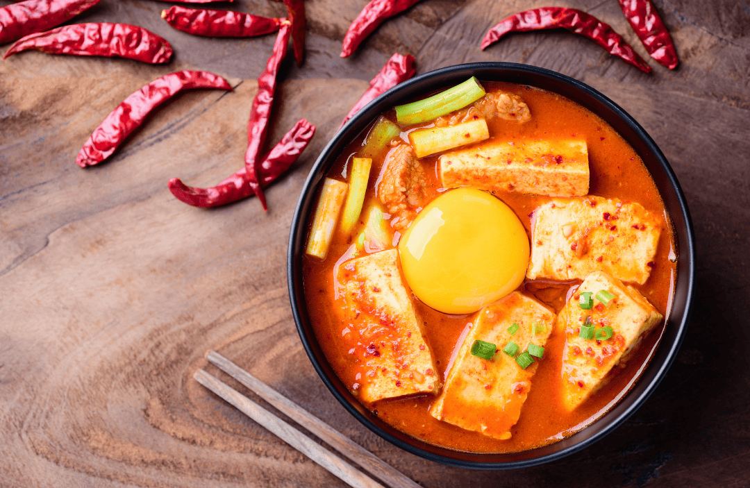 A bowl of spicy Korean tofu stew (soondubu jjigae) with soft tofu, vegetables, and a raw egg yolk on top, surrounded by dried red chili peppers on a wooden background.