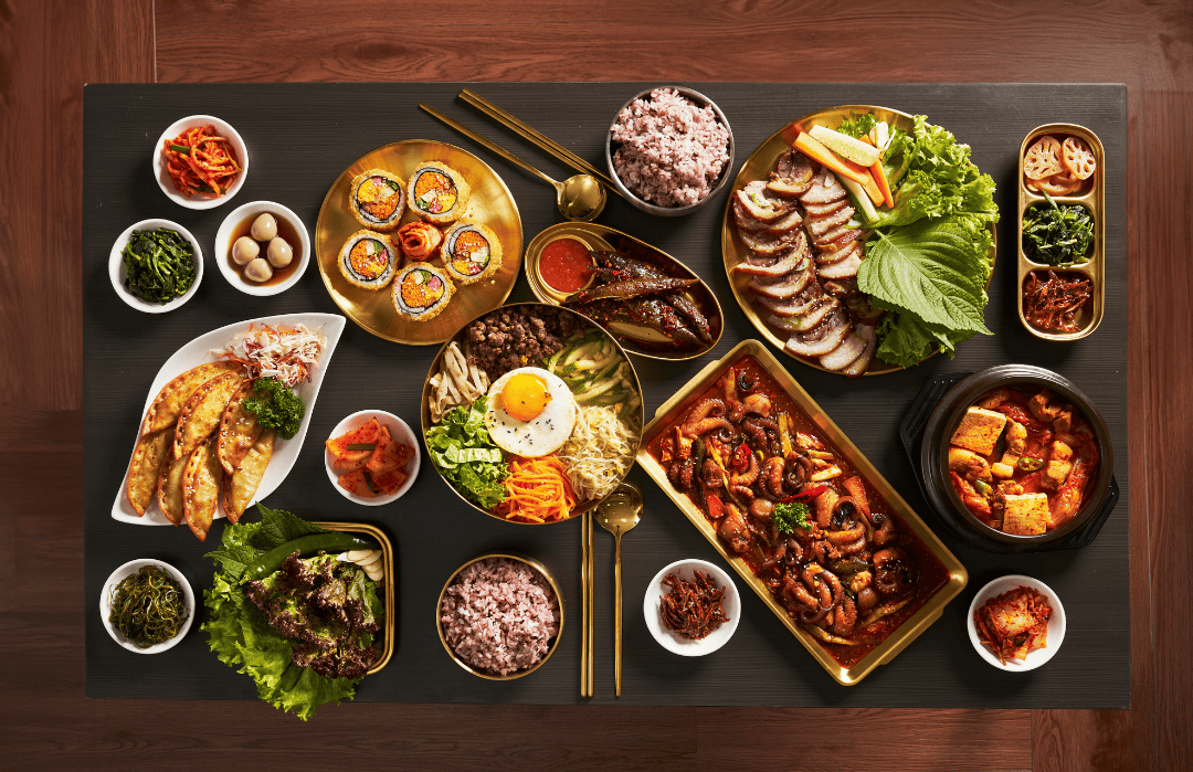 A traditional Korean table setting featuring a variety of dishes including gimbap, bibimbap, grilled meat, tofu stew, and assorted side dishes on a black table with gold utensils.