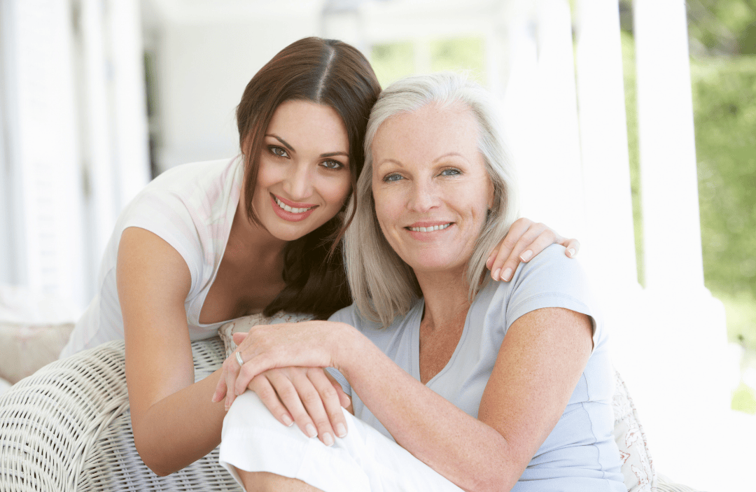 A mother and adult daughter embracing and smiling together while sitting on a porch.