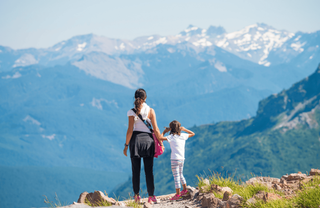  A mother and daughter hiking together in the mountains, facing scenic snow-capped peaks.